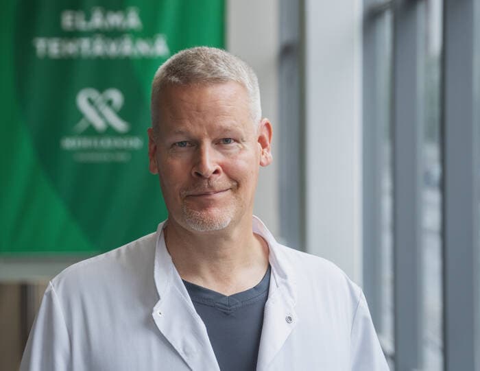 Mehiläisen ortopedi päällään valkoinen lääkärin takki katsoo kameraan hymyillen. Taustalla Mehiläisen vihreä mainoskyltti.