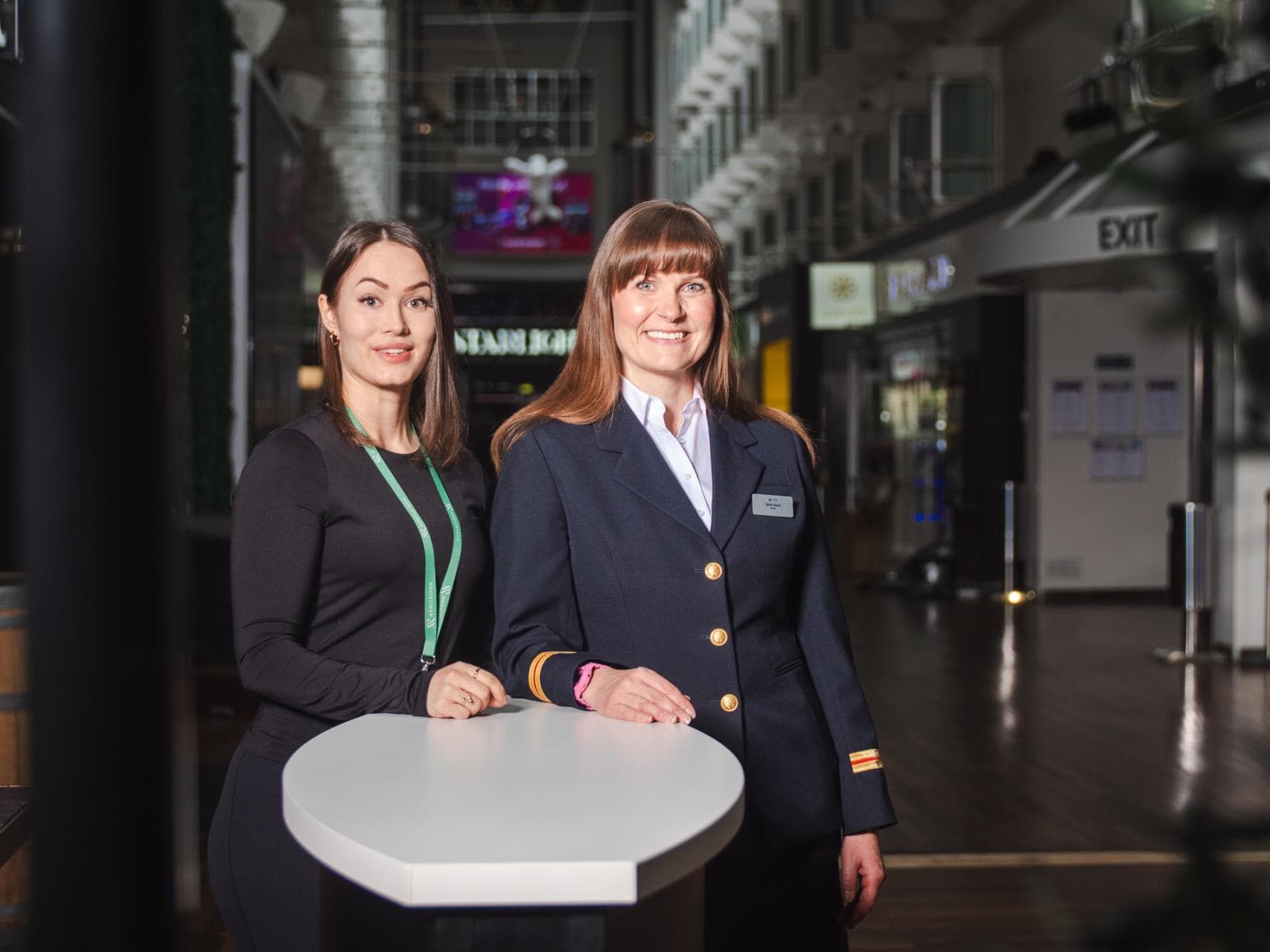 Kaksi hymyilevää naista seisoo valkoisen pöydän takana. Vasemmalla oleva nainen on pukeutunut mustaan asuun ja hänellä on vihreä kaulanauha. Oikealla oleva nainen on pukeutunut tummansiniseen virkapukuun, jossa on kultaiset napit ja hihamerkit. Taustalla näkyy laivan sisätiloja. Kuvassa Tallink Siljan laivasairaanhoitaja Terhi Vuori ja Mehiläisen ravitsemusterapeutti Johanna Räsänen.