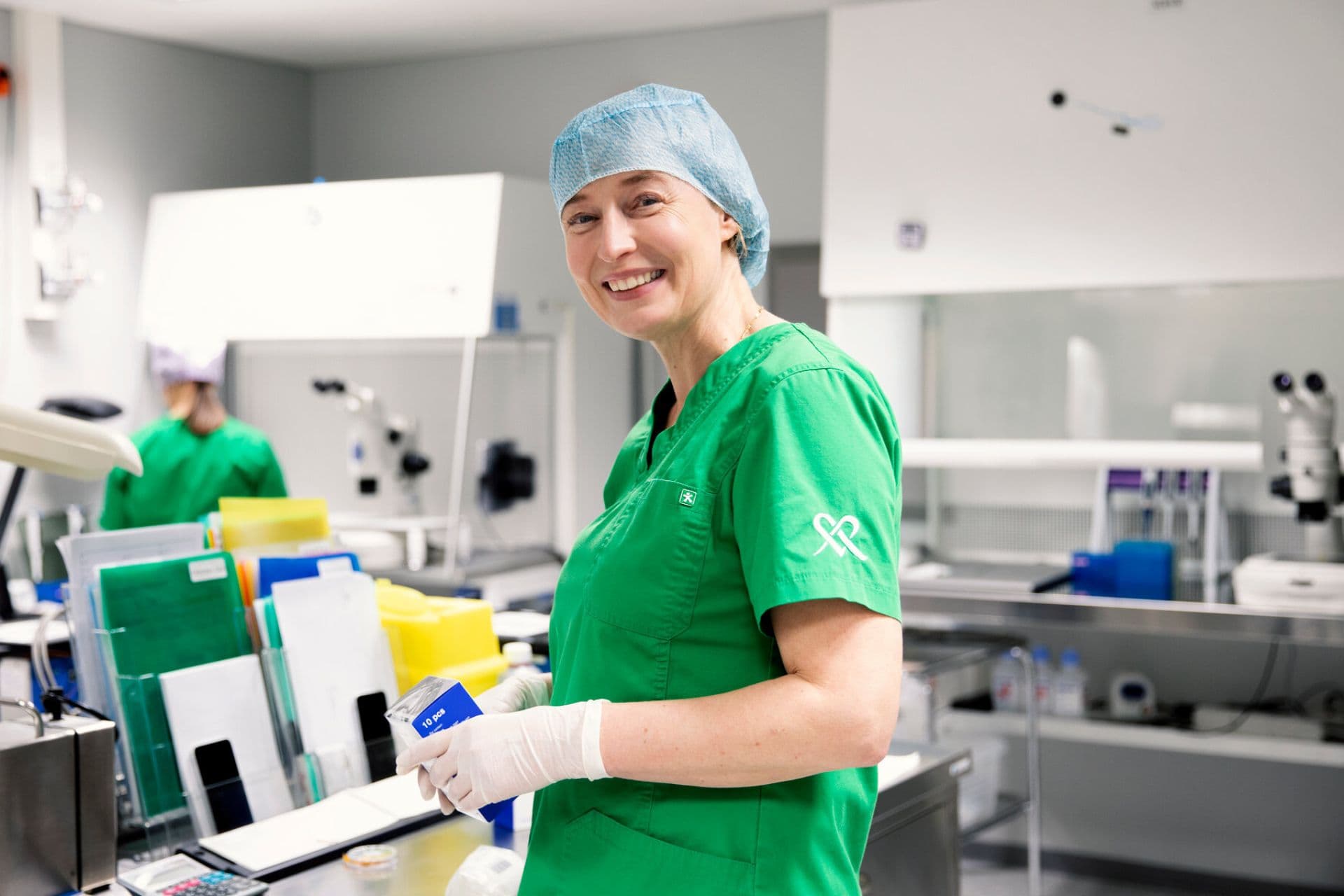 Lääkäri hymyilee kameralle laboratoriossa. Hänellä on yllään vihreä lääkärintakki, valkoiset hanskat ja sininen hiussuoja. Taustalla näkyy laboratoriolaitteita ja toinen henkilö työskentelemässä.