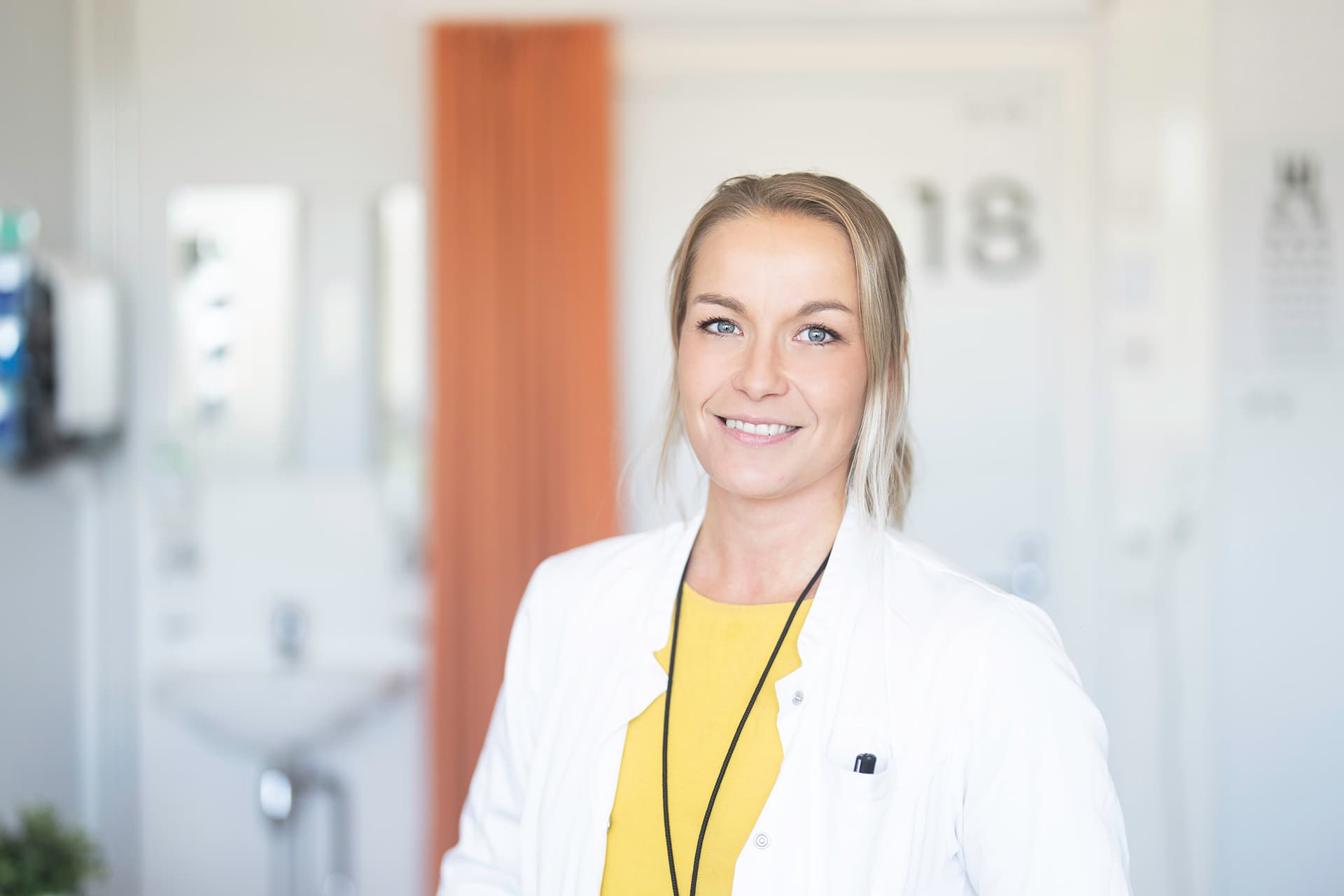 Mehiläisen korvalääkäri seisoo iloisena vastaanottohuoneen edessä lääkärintakkiin pukeutuneena.