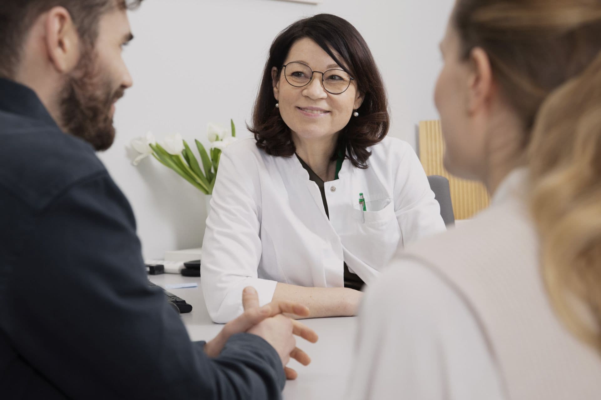 The physician is having a discussion with a couple at the practice. They are sitting behind the desk and looking towards the couple. Of the couple, only the man on the left and the woman on the right are partially visible.