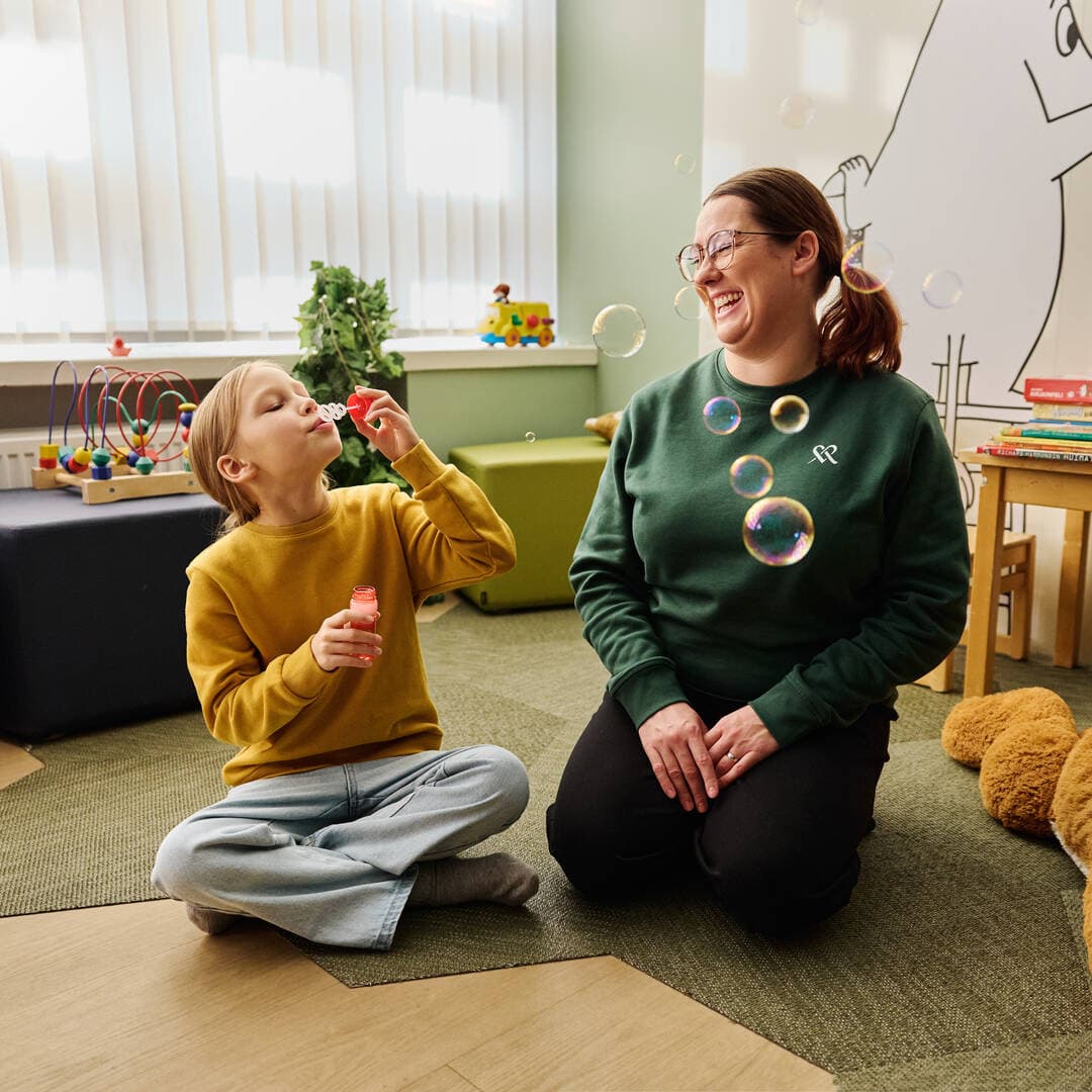 The therapist sits on the floor with the child and smiles as the child blows soap bubbles